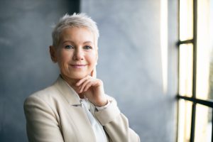 Elegant Middle Aged Businesswoman With Stylish Pixie Hair Standing By Window In Office, Touching Face, Thinking About Business Issues, Looking At Camera With Confident Smile. Success, Career And Job